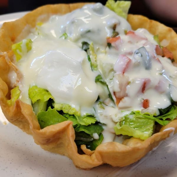 A taco salad in a crispy fried tortilla bowl, topped with lettuce, tomatoes, and white sauce.