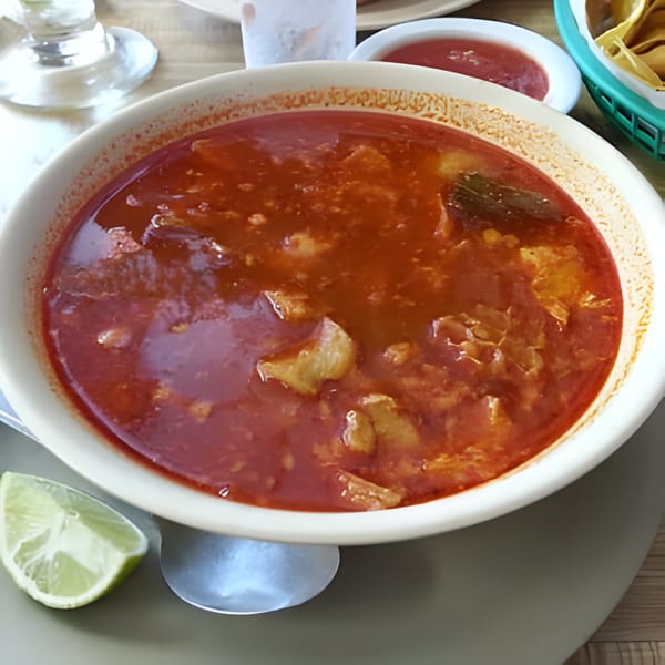 A bowl of Mi Rinconcito's red menudo soup with meat, a lime wedge, and salsa.