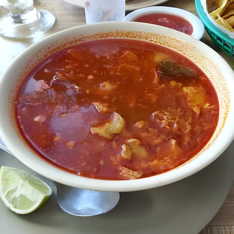 A bowl of Mi Rinconcito's red menudo soup with meat, a lime wedge, and salsa.